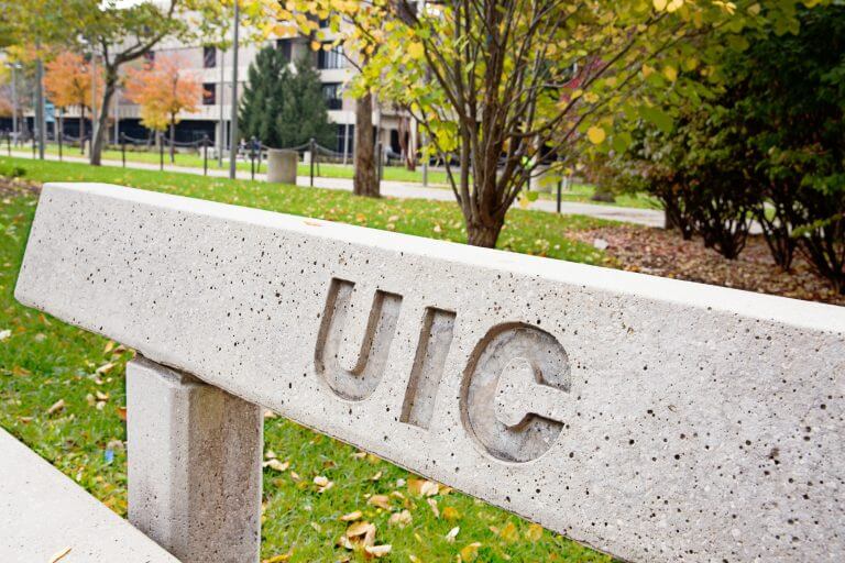 Cement park bench with UIC engraved, in front of grassy/tree area on campus