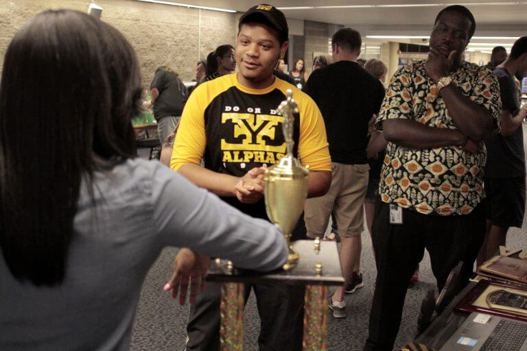 Male student of color wearing Greek-letter shirt talking to someone leaning on large trophy