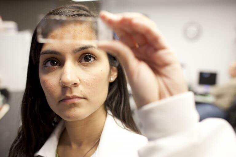 Hispanic researcher in white lab coat looks at specimen slide she is holding up