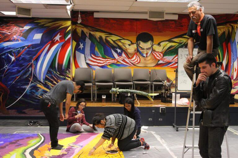 several students work on colorful mural on ground in office space with colorful diverse mural on wall