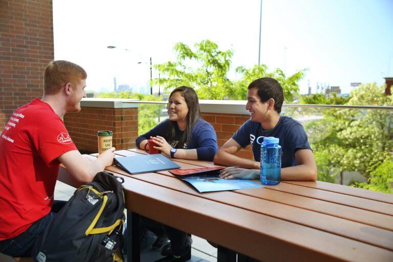two male and one female student sit at brown table outdoors smiling and talking