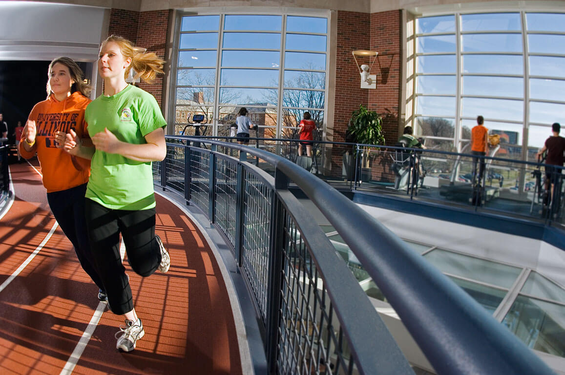 two women jogging on track inside rec center, with others on stair climbing machines in background