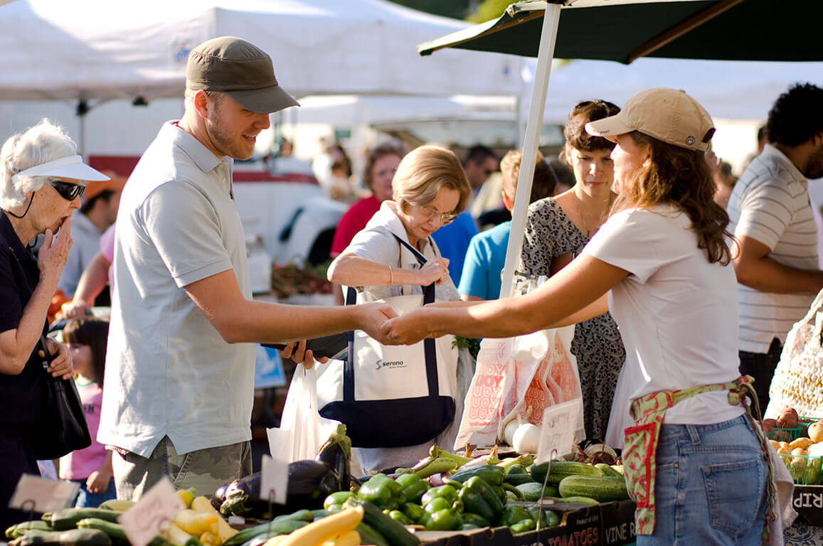 Man in hat purchasing food at an outdoor farmers market from female vendor