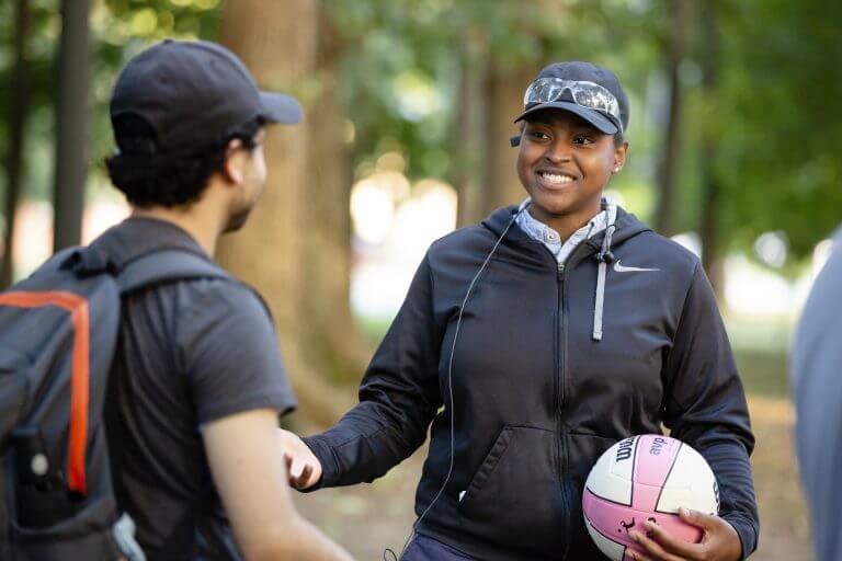 black student in ball cap smiling, wearing hoodie jacket, holding volleyball talking to another student in hat