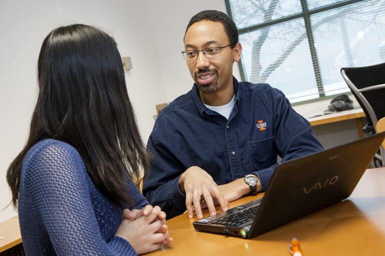 Male of color with glasses wearing ILLINOIS button down-shirt talking to female student with laptop on table in office
