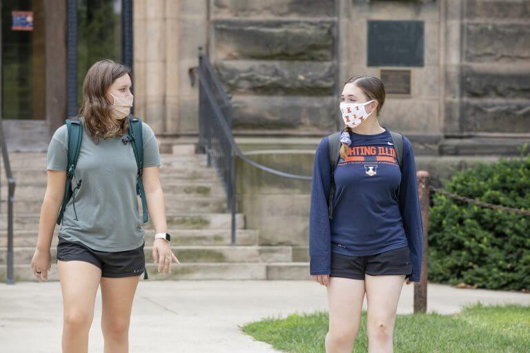 female students in mask leaving campus building with backpacks, one with Illinois shirt