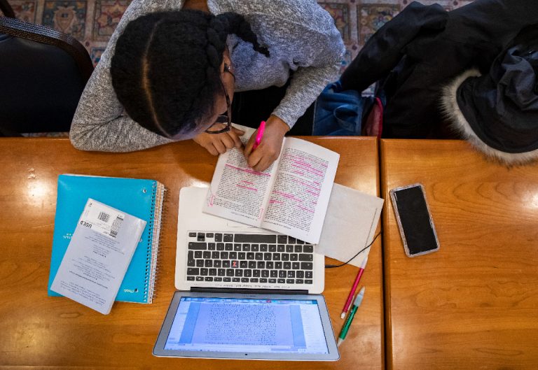 overhead view of female student highlighting passages in a book on a table with laptop