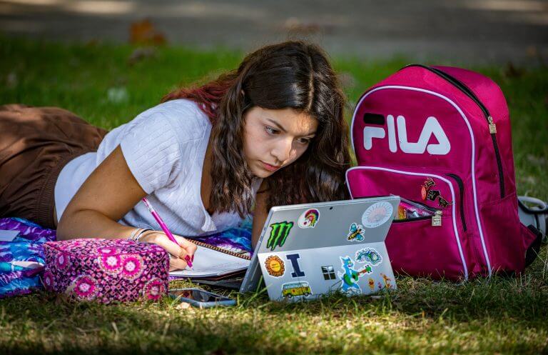 Hispanic female student lying on stomach on grass, studying with pink backpack