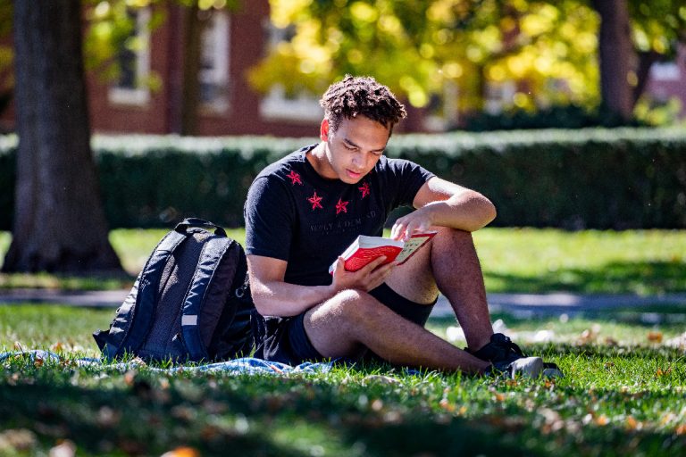 Male student of color reading on lawn on campus in shorts and T-shirt, with backpack on ground