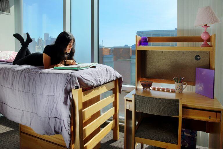female student lying on stomach on lofted bed studying in dorm room with Chicago view out of window