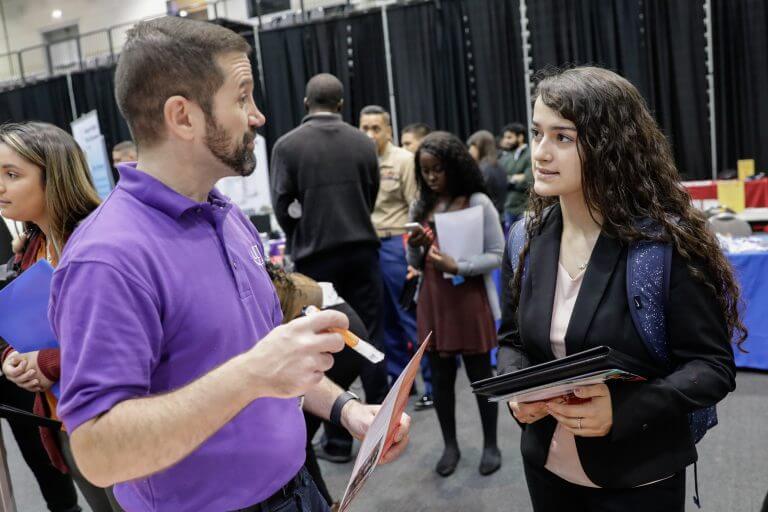 101719_Diversity-Career-Fair_JC0010-2-768x512.jpg bearded male in purple polo with pen and flyer talks to female student at a career fair