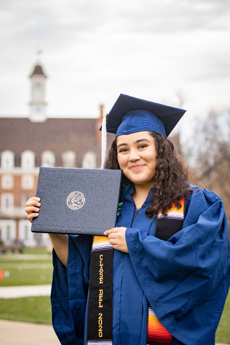 Hispanic female in cap and gown holding up diploma in front of Illini Union